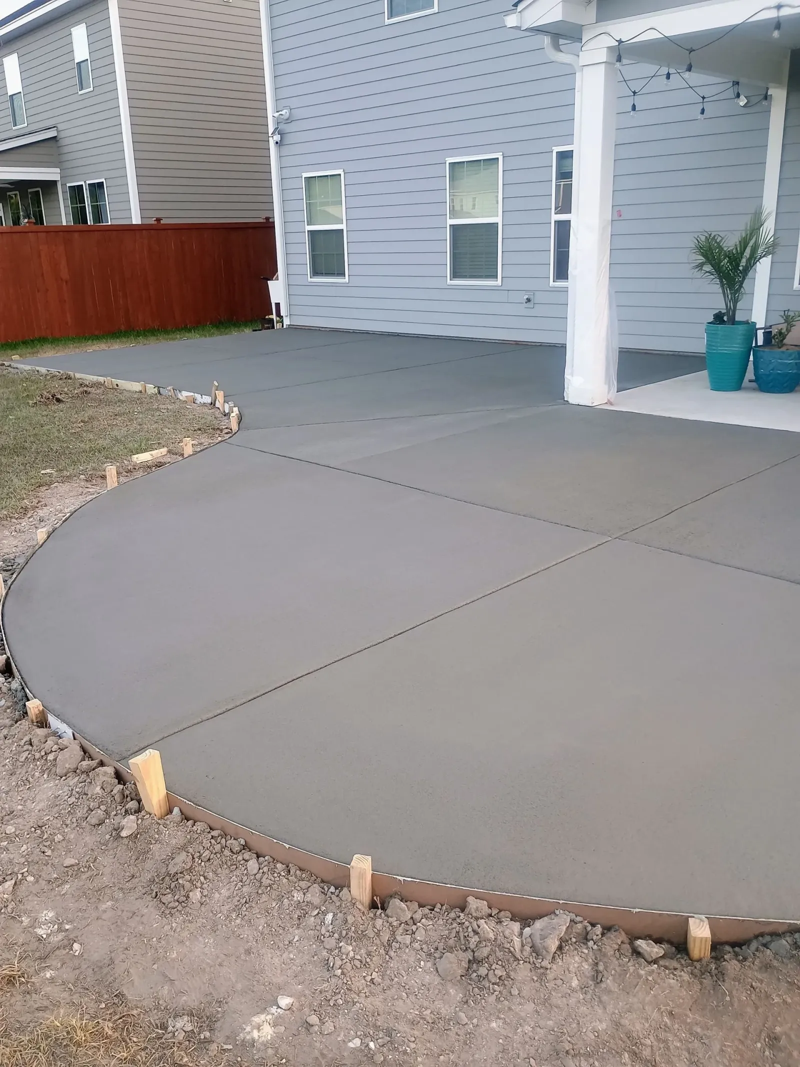 Fresh concrete driveway leading to a gray house with fenced sides and dirt along the edges