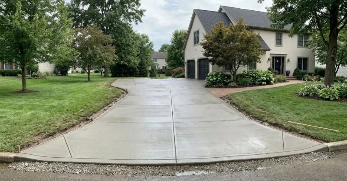 Fresh concrete driveway leading to a gray house with fenced sides and dirt along the edges